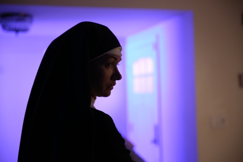 A nun stands in profile in a white room. The room behind her is bathed in a purple light.